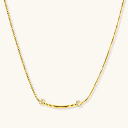 Delicate gold-plated necklace with curved bar pendant and crystal accents on snake chain against cream background.